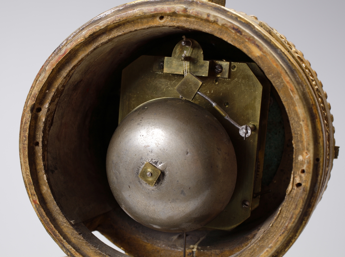 Bordspendyl / Bordsstudsare.
Vit emaljerad urtavla försedd med arabiska siffror i en rund, guldbronserad överdel av trä, som vilar på tre snidade delfiner. Högst upp en guldbronserad urna. 
Cylindrisk fot av trä, målad i rödbrun marmorimitation med guldbronserade partier. Tillhörande urnyckel av järn.