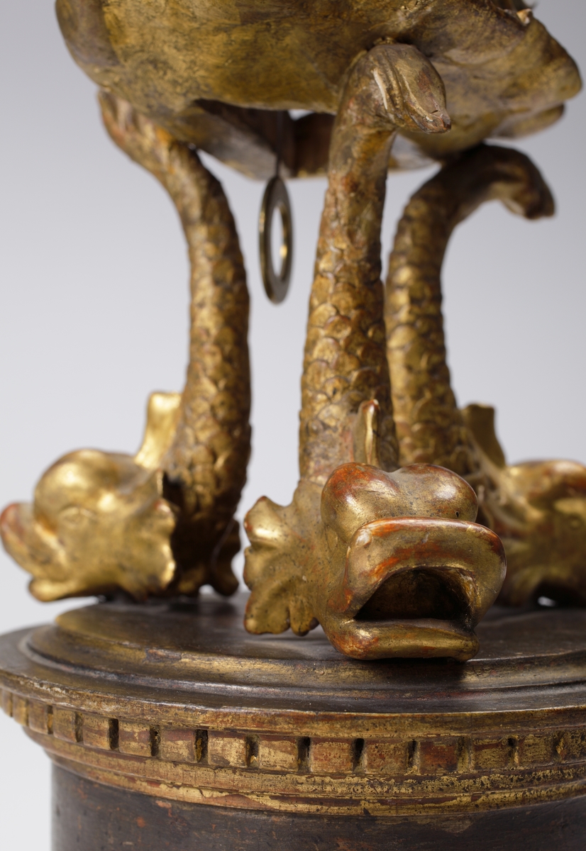 Bordspendyl / Bordsstudsare.
Vit emaljerad urtavla försedd med arabiska siffror i en rund, guldbronserad överdel av trä, som vilar på tre snidade delfiner. Högst upp en guldbronserad urna. 
Cylindrisk fot av trä, målad i rödbrun marmorimitation med guldbronserade partier. Tillhörande urnyckel av järn.