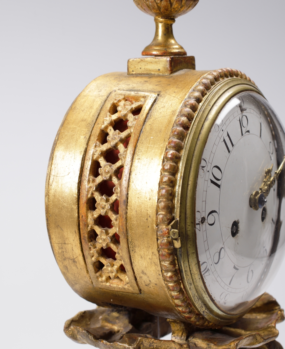 Bordspendyl / Bordsstudsare.
Vit emaljerad urtavla försedd med arabiska siffror i en rund, guldbronserad överdel av trä, som vilar på tre snidade delfiner. Högst upp en guldbronserad urna. 
Cylindrisk fot av trä, målad i rödbrun marmorimitation med guldbronserade partier. Tillhörande urnyckel av järn.