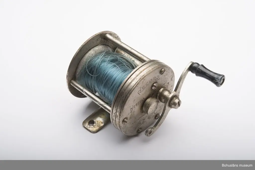 Skall fästas på ett spinnspö.
Två runda metallplattor vilka är sammansatta parallellt på 4,5 cm avstånd med fem stycken metallstavar. På den ena staven är det en an-
ordning som får fisklinan att fördelas jämt på rullen. Vev på ena sidan med ett litet handtag i svart- och gråmelerad ureaplats.
Ursprungligen har det varit två handtag.
Text på den ena metallplattan:"RECORD NO 1550 A.-B. -URFABRIKEN - SVÄNGSTA". På den andra plattan är det ristat:"Bertil Becker" samt
något oläsligt. Blå lina av polyamidfiber upplindad på rullen. Metallplattan undertill vilket är en fästanordning för att få fast rullen
vid ett spö. På plattan står det:"MADE IN SWEDEN".
Linan är troligen från 1950-talet.