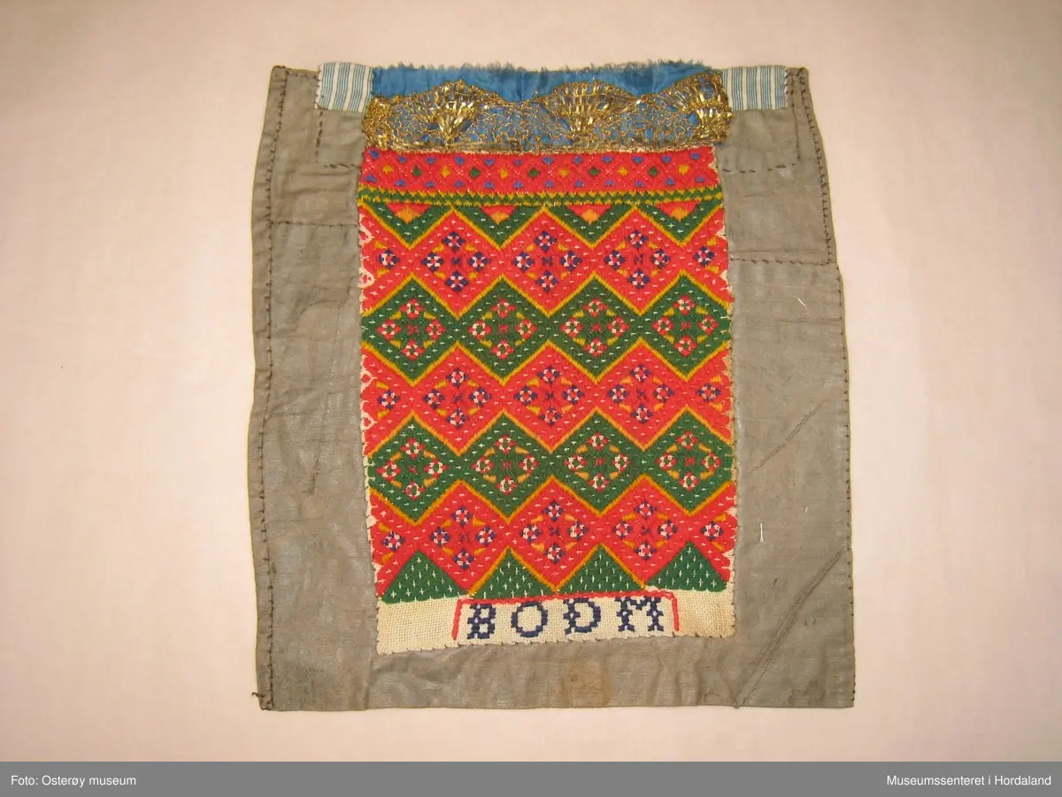 Brodert med ullgarn i plattsaum. Alt er handlaga. Hovudmotiv er små åttebladroser i border. Oppe er det påsydd eit blått silkeband med eit breitt gullband (i forgylt sølvtråd) utanpå silkebandet. Alt er montert på eit tynt, vokset bomullslerret. Oppe på baksida er det eit ekstra bomullstoff (blåstripet). Bokstavane BODM er broderte inn nede på bringeduken i kross-sting.