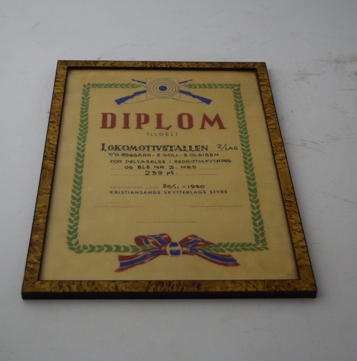 To skytevåpen i kryss med en blink over, sentralt oppe. Bladranke som ramme rundt skriften med en sløyfe på midten nederst.