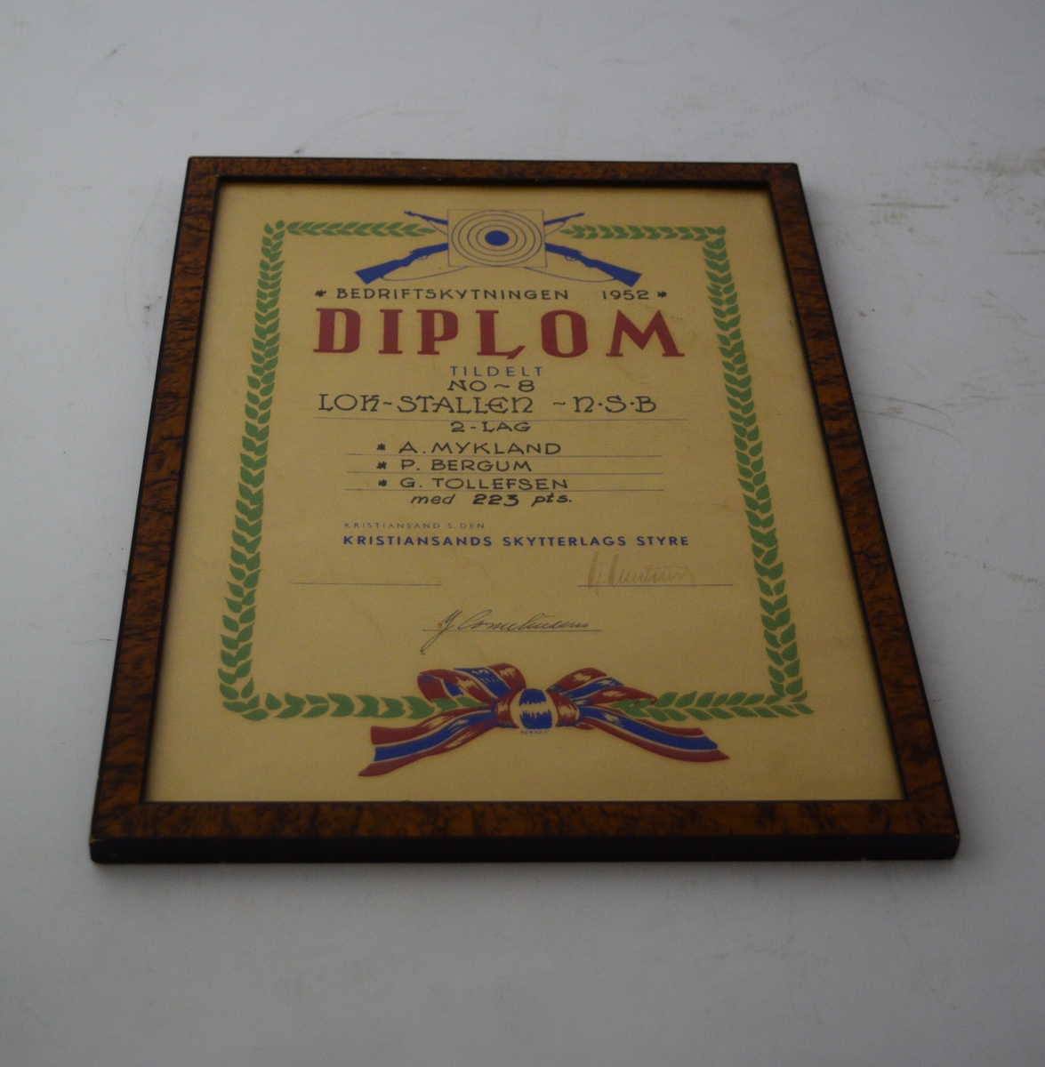 To skytevåpen i kryss med en blink over, sentralt oppe. Bladranke som ramme rundt skriften med en sløyfe på midten nederst.