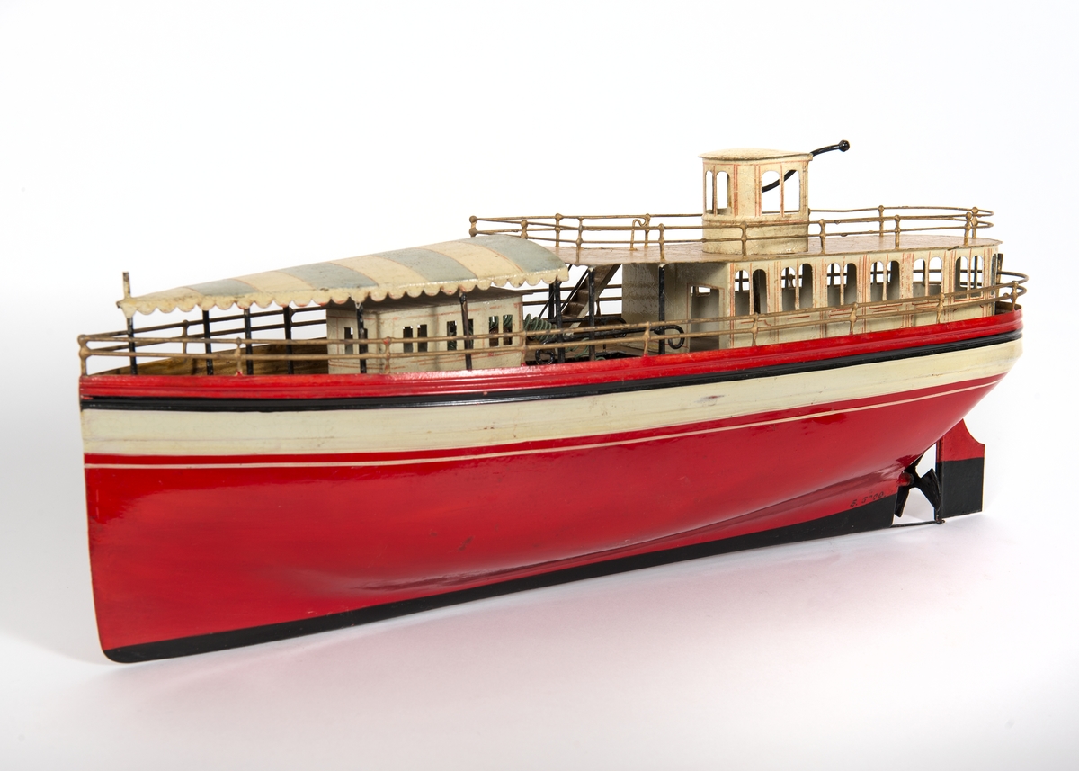 Fartygsmodell. Leksaksbåt av plåt. Rödmålat skrov med svartvitt bälte över vattenlinjen. Rak förstäv, häckakter, roder och en trebladig propeller. Förligt däckshus överspänt med "suntält" av plåt, akterut passagerarsalong, navigationshytt. Propellermaskineriet består av en tvåpolig elektrisk motor driven av ett De Lanche element. Gråmålat skrå.