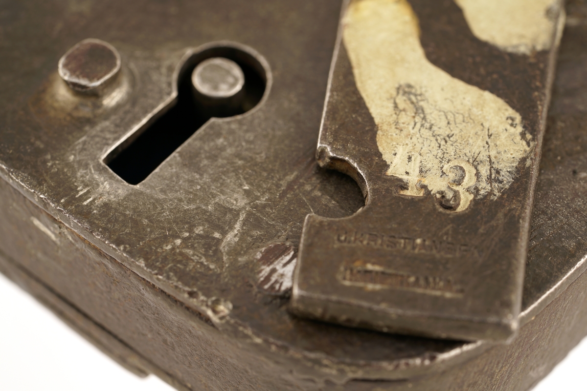 Jern, firkantet, svingbart lokk over nøkkelhullet.
Nøkkel, jern, firkantet skjegg, gjennomboret.
Papirlapp knyttet fast i nøkkel med snor.