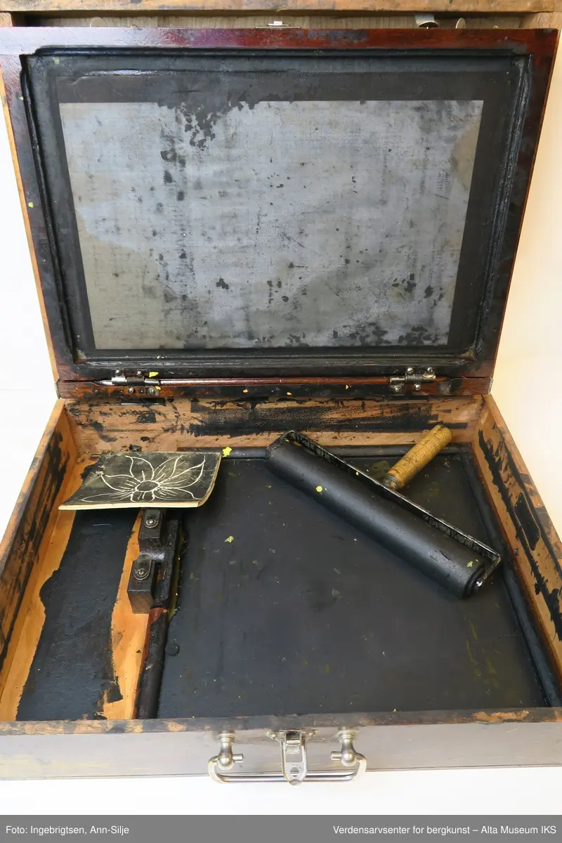 Trekoffert med et lite metallhåndtak. Inn i kofferten er det sverterull, plater, et rutet ark og en sjablong med motiv av en blomst. Både platene og det inngraverte stykket har et lag av inntørket sverte.