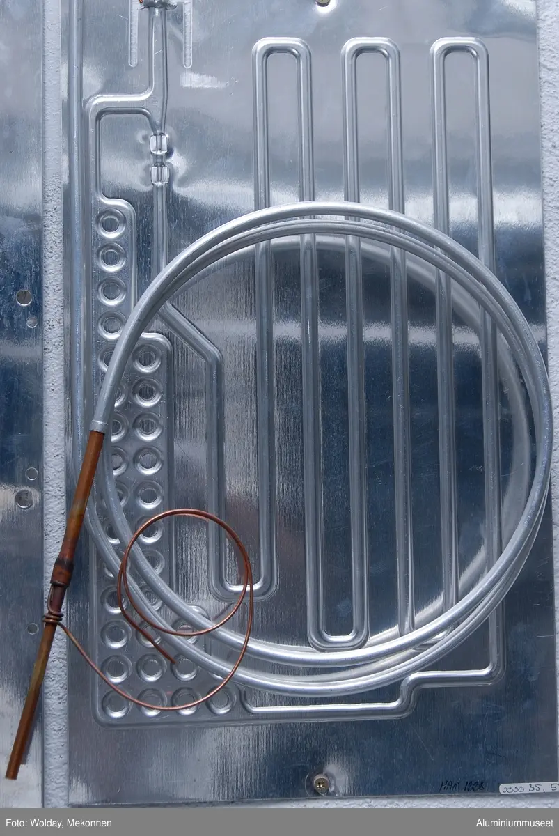 Teknikk: En aluminium-plate med riktig bredde, med kortere lengde og med større tykkelse enn den ferdige fordamperen ble med siketrykk påført mønsteret til fordamperen, se bilde nr. 31. Mønsteret var som platen riktig på bredden, men lengden var redusert. En plate med samme dimensjon ble lagt oppå den første platen og disse to ble varmvalset sammen. Platene ble etterpå finvalset slik at lengden ble 1,4 ganger lengre og da var også mønsteret innvendig valset til riktig dimensjon. Siketrykkmønsteret var skåret riktig på alle breddemål, mens lengdemålene var redusert med 1,4. På bildet kan man se at kanalbredden på 10 mm. er riktig i lengderetning, men vesentlig redusert i bredderetning. Etter finvalsing ble alle kanalene valset til 10 mm. Platene ble videre glødd i valseverket og så sendt til Fordamperavdelingen for videre produksjon.
I Fordamperavdelingen ble platene som nå var helt plane blåst opp i en motholdspresse slik at alle kanalene fikk riktig høyde. Panelene ble så formatklippet, lokket, stanset, rør ble påsveiset, trykkprøvet og lakkert. 
Form: Firkantet form
