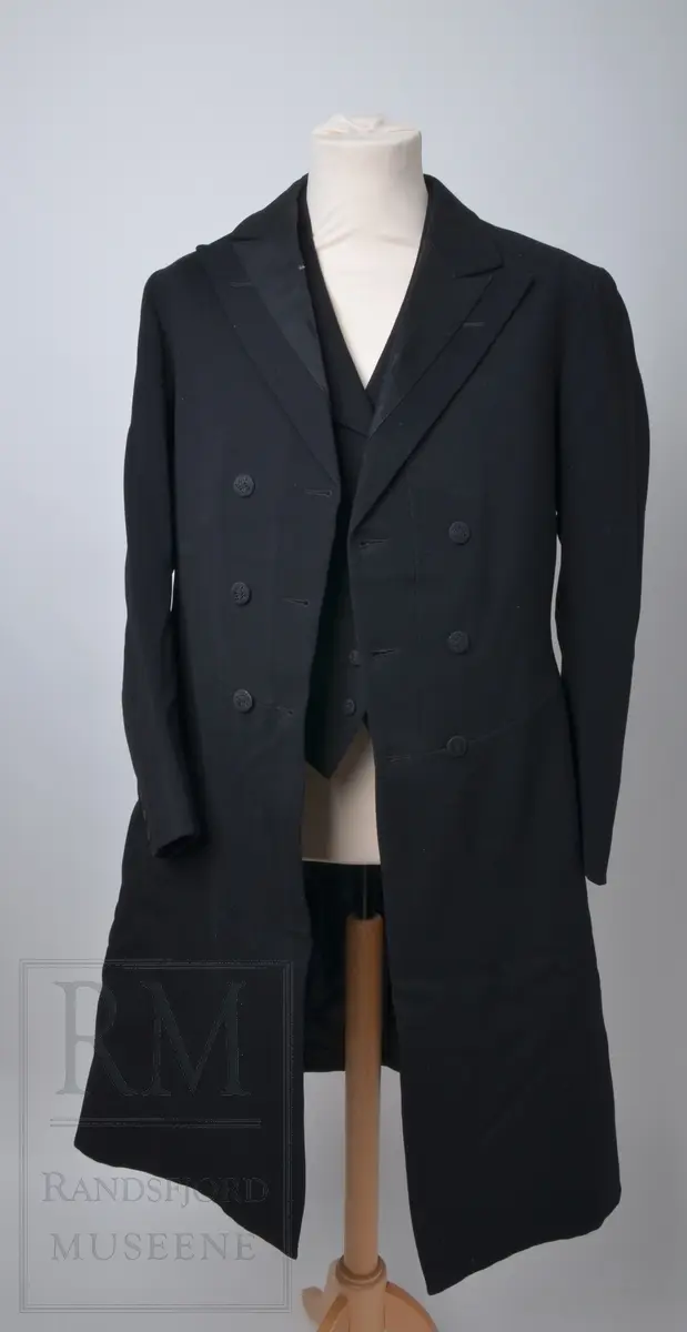 Tredelt dress med hvite hansker. Frakk av livkjoletype, kalt bonjour. Vesten har slag som er fôret med svart silkefôr på innsiden og har seks trekte knapper. Bunnfargen på fôret er hvit med brune og grå striper.