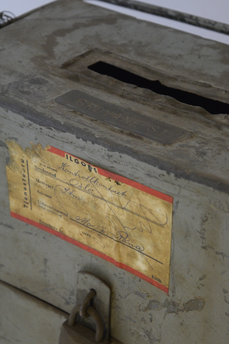 Firkantet kasse, smal rektangulær åpning i lokket. Luke i front nede, lukket med hengsle og hengelås. Hank festet på sidene. Papiretikett klistret på foran.