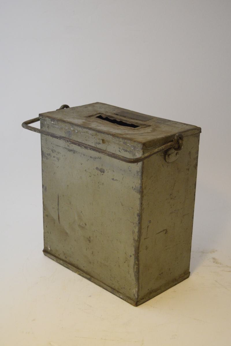 Firkantet kasse, smal rektangulær åpning i lokket. Luke i front nede, lukket med hengsle og hengelås. Hank festet på sidene. Papiretikett klistret på foran.