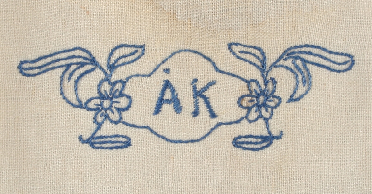 Ermlaus kjole av bærestykke og skjørt. Lomme midt framme. Handbroderi i kontursting. Blå broderitråd tredd i maskinsaumen lagar dekor rundt hals, skulder og lomme. Maskinvovne pynteborder. Tre trykk-knappar lukkar bærestykket bak. Monogram og blomar på bærestykke; ÅK = Åse Kristin. Fugl på grein på lomme. To kattar framme på skjørtet. To høner bak på skjørtet.