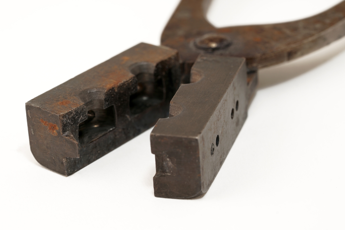 Plombestøpetang av jern m/ treskaft.
Utstyrt med fire hull. De to på oversiden var for flytende bly. De to på siden var til bruk for en spesiell
"gaffel", som skulle lage hull i plombene for forseglingstråden.