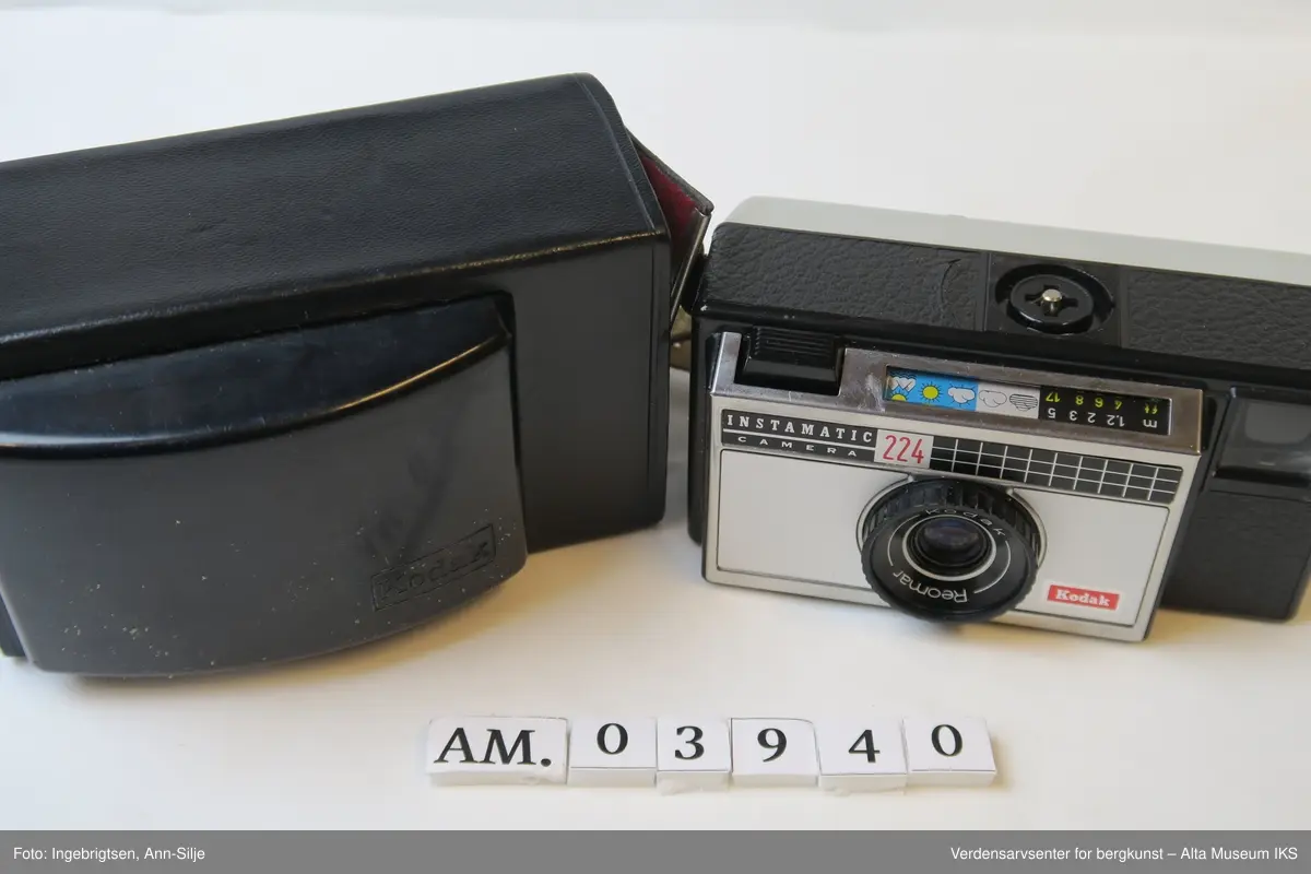 Fotoapparat i rektangulær form. Den er festet i et læretui med rødt tøystoff på innsiden. Inn i apparatet er det en filmrull av merket Fuji. På toppen av apparatet, ved siden av utløseren, er det symboler av vær og landskap.
