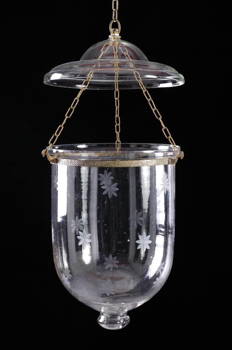 Ljusampel av glas.
Cylindrisk form med rund botten, avslutad med knopp. Dekor av slipade stjärnor (blommor?). Runt ampelns övre kant löper ett band av graverad mässing, med tre gjutna kedjehållare utformade som sjöhästar. Över ampeln ett profilerat, runt ljustak av glas. Kedjor av mässing.