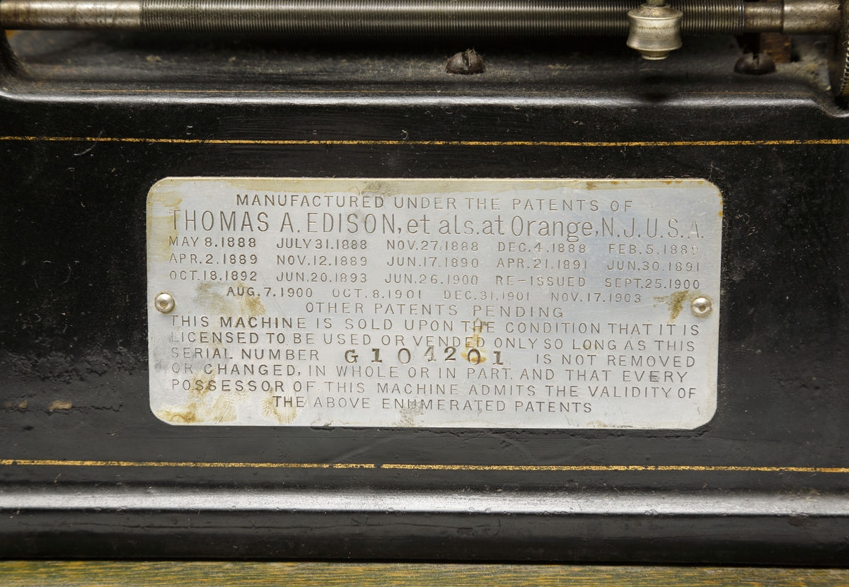 Fonograf, med förvaringslåda. 
Rektangulär, svartlackerad metallåda med musikinstrumentet ovanpå. Dessutom en förnicklad (?) tratt. Vrides upp med nyckel på kortsidan. 
Förvaringslåda av fernissat trä med handtag och påmålad text: "Edison, GEM, Phonograph".
