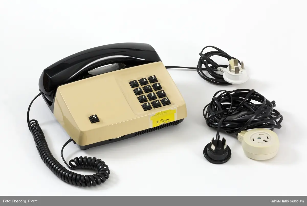 KLM 46164:1-2. Telefon, telefonapparat, knapptelefon. Av beige och svart plast. Tillverkare: Telia, Televerket. Modell: Diavox. I rektangulär plan form med svart knappsats till höger. Vid fronten en gul etikett med angiven adress. Medföljer svart elsladd och förlängningssladd med vit telefonkontakt.