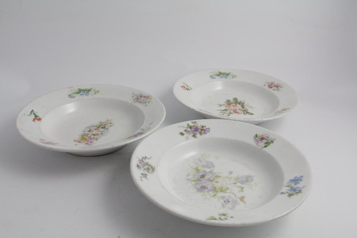Runde dype tallerkener, tre stykker. Hvit bunn, polykrom blomstermotiv (naturalistisk) trykket på. Blomsterbukett på speil, fem mindre buketter på fane. Hver tallerken er forskjellig. Merket med produsentlogo under.