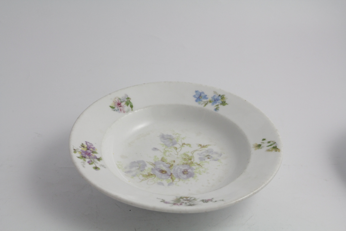 Runde dype tallerkener, tre stykker. Hvit bunn, polykrom blomstermotiv (naturalistisk) trykket på. Blomsterbukett på speil, fem mindre buketter på fane. Hver tallerken er forskjellig. Merket med produsentlogo under.