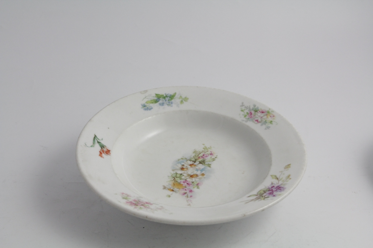 Runde dype tallerkener, tre stykker. Hvit bunn, polykrom blomstermotiv (naturalistisk) trykket på. Blomsterbukett på speil, fem mindre buketter på fane. Hver tallerken er forskjellig. Merket med produsentlogo under.
