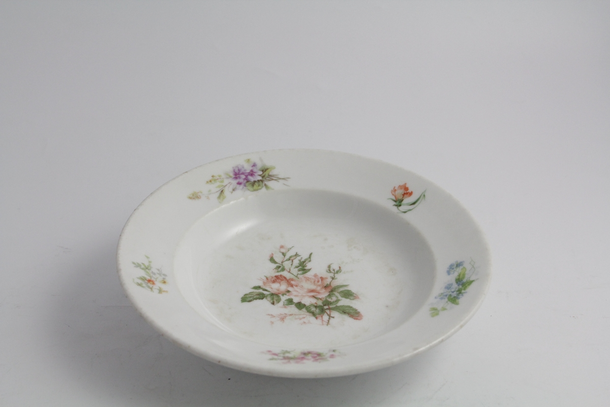 Runde dype tallerkener, tre stykker. Hvit bunn, polykrom blomstermotiv (naturalistisk) trykket på. Blomsterbukett på speil, fem mindre buketter på fane. Hver tallerken er forskjellig. Merket med produsentlogo under.