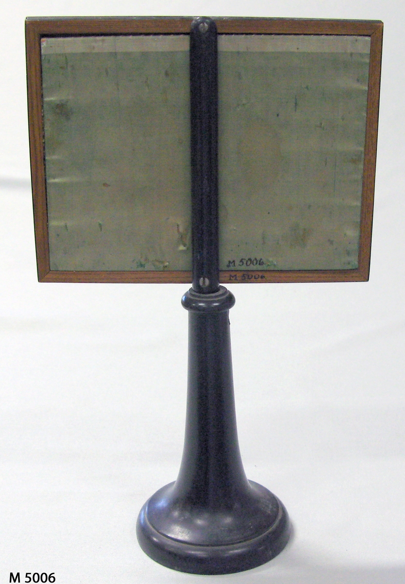 Ljusskydd.
Svart träram på svarvad träfot. I ramen körssömsbroderi å stramalj i 
färgerna gråsvart och vitt. Motiv: kyrka. Baksidan täckt med grönt siden (japonne).
Ramens storlek: 19,5 x 15 cm. Fotens höjd: 16,5 cm. 
Diameter nedtill 10 cm.
Funktion: Ljusskydd