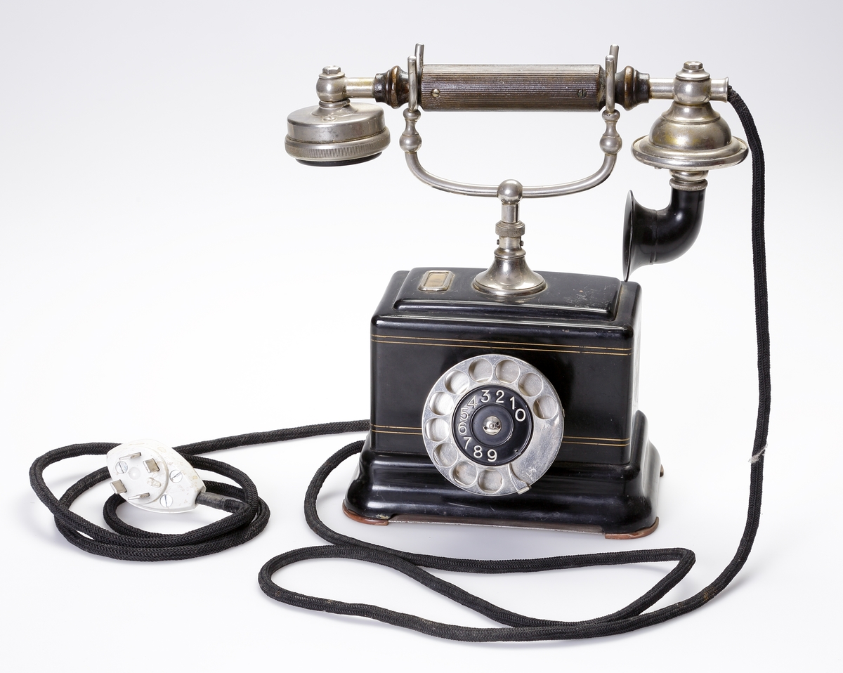 Telefon, bordsmodell. 
Rektangulär, svart bakelitmodell med detaljer av nickel. Telefonlur med svart tygsladd.
