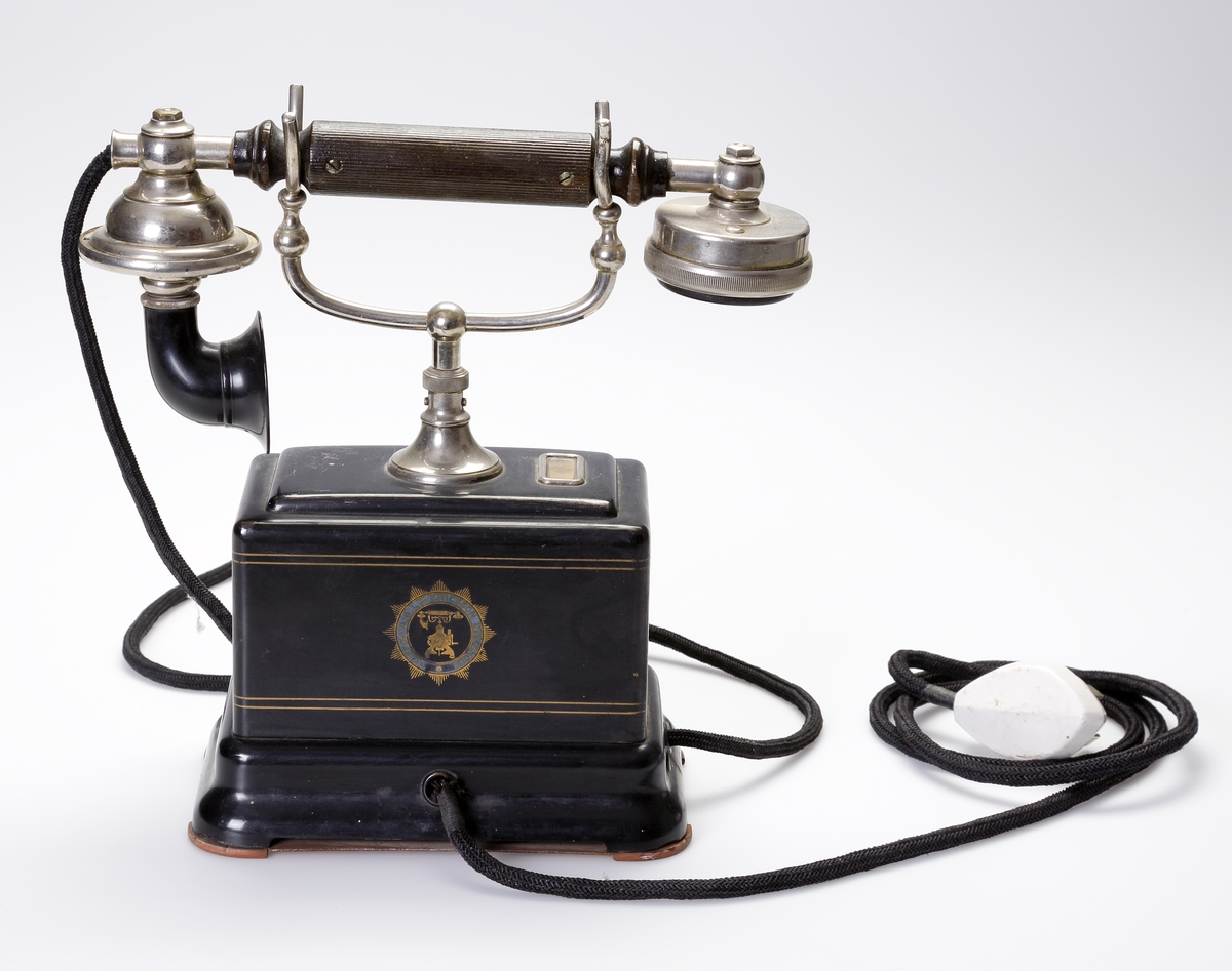 Telefon, bordsmodell. 
Rektangulär, svart bakelitmodell med detaljer av nickel. Telefonlur med svart tygsladd.