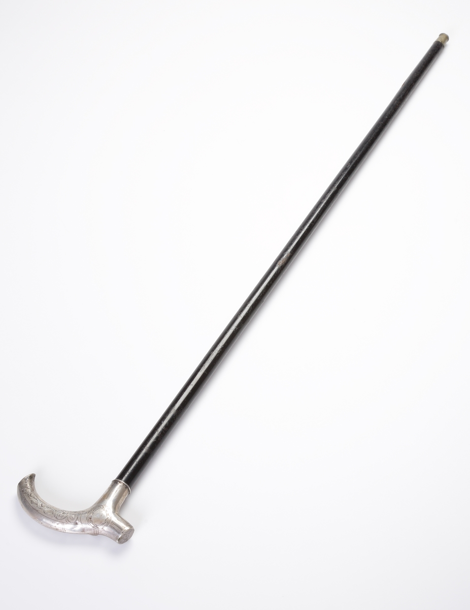Käpp, s.k. spatserkäpp.
Svarvat, svartlackerat trä. Krycka och skoning av silver. Kryckan försedd med gravyr i nyempire och monogram: "A.A.". Stämplar (något otydliga) längs nedre kanten av kryckan.