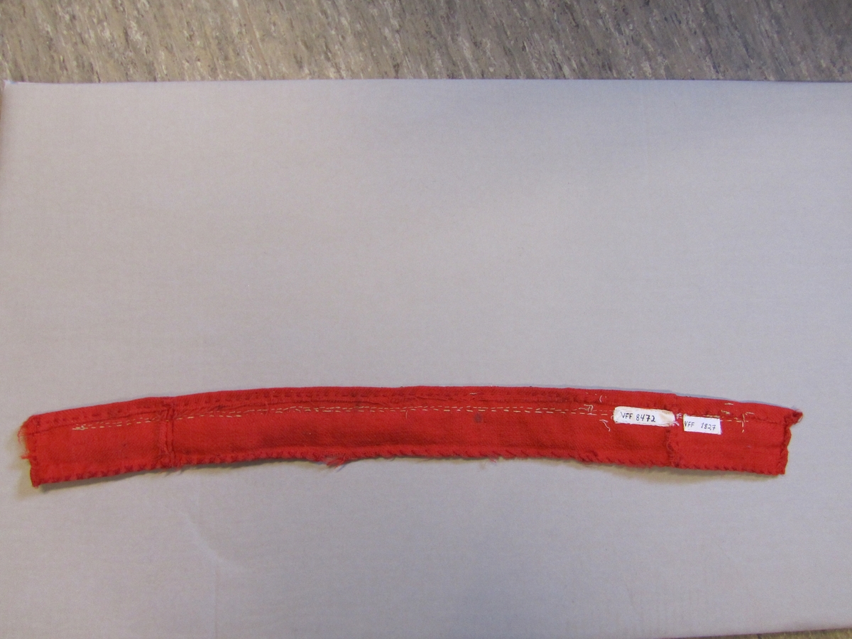 Perleband.  Eit raudt kypertvove ullband som er bretta inn på båe kortsidene og eine langsida. Nedsydd med lange forsting frå retta med raud tjukk ulltråd.  Den andre langsida er kasta over med same type ulltråd.  Påsydd eit perla band med glassperler og ei vove metallbore.