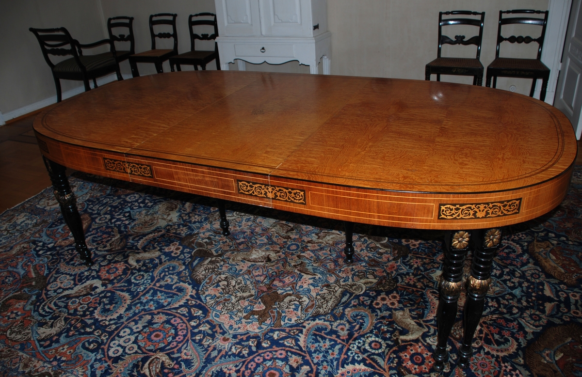 Uttrekkbart spisebord laget av snekkermesterfirma Knag i Bergen 1939. Lys finér, med intarsia på sidene. Benene er dreiede, sorte med gulldetaljer.