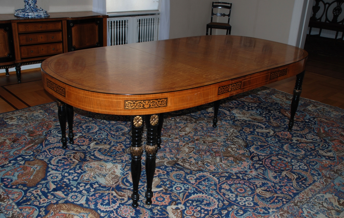 Uttrekkbart spisebord laget av snekkermesterfirma Knag i Bergen 1939. Lys finér, med intarsia på sidene. Benene er dreiede, sorte med gulldetaljer.