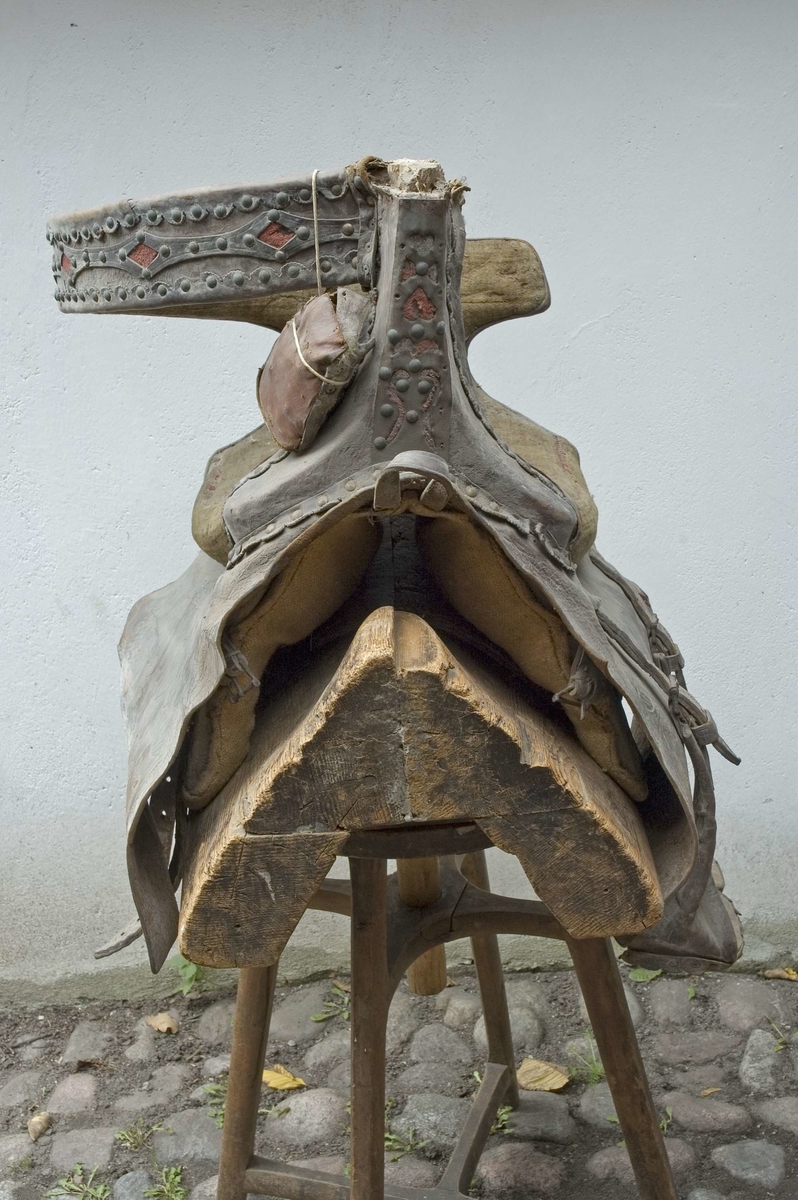 Brudsadel, handgjord. Modell där den ridande färdas sidledes, med fötterna på samma sida på ett fotstöd. Försedd med ryggstöd.
Stomme av trä. Sitsen, sido- och ryggstöden stoppade och klädda med grön ylleplysch, nersydd i mönster med röd tråd, troligen av lin. På utsidan är stöden klädda med brunt skinn, plåt och nitar. Både plåt och skinn har genombrutna partier, med underliggande rött kläde. Märkningen GTDR (osäker) samt 1809 är utskuret i plåt med rött kläde under. Detta är placerat på bakre sidostödet. På båda sidorna hänger stycken av läder med rundade hörn, med präglat mönster som liknar en blomsteruppsättning av barocktyp. Fotstödet hänger på vänster sida, i justerbara remmar av läder med söljor av metall. Trästommen är också stoppad ned mot hästryggen, och klädd med säckväv av jute. Det finns också remmar för fastsättning av sadeln, men de är korta och måste skarvas. Allt trä klätt med målad säckväv.