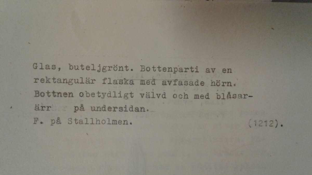 Underdel av en åttkantigt, rektangulär, buteljgrön glasflaska. Bottnen obetydligt välvd och med blåsarärr på undersidan. Enligt den ursprungliga fyndjournalen skall det ursprungligen ha funnits 3 fragment.