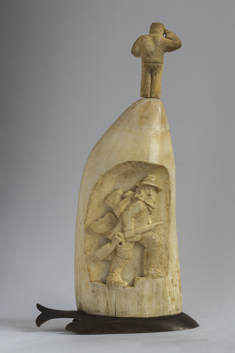 Hvaltannskulptur på plint av messing formet som en hval. Utskåret jegermotiv i hvaltannen.