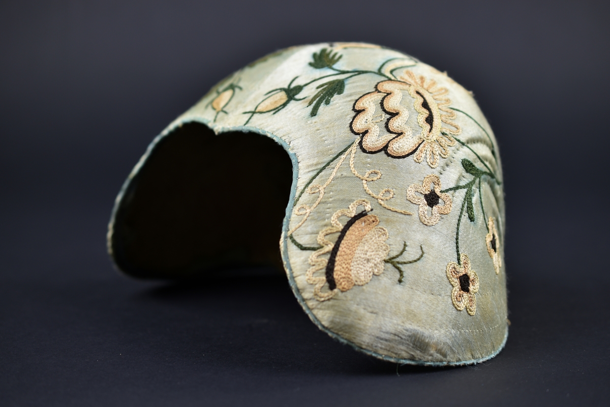 Bindmössa av ljusblått siden dekorerad med broderade blommor och blad. Foder i oblekt linnetyg.