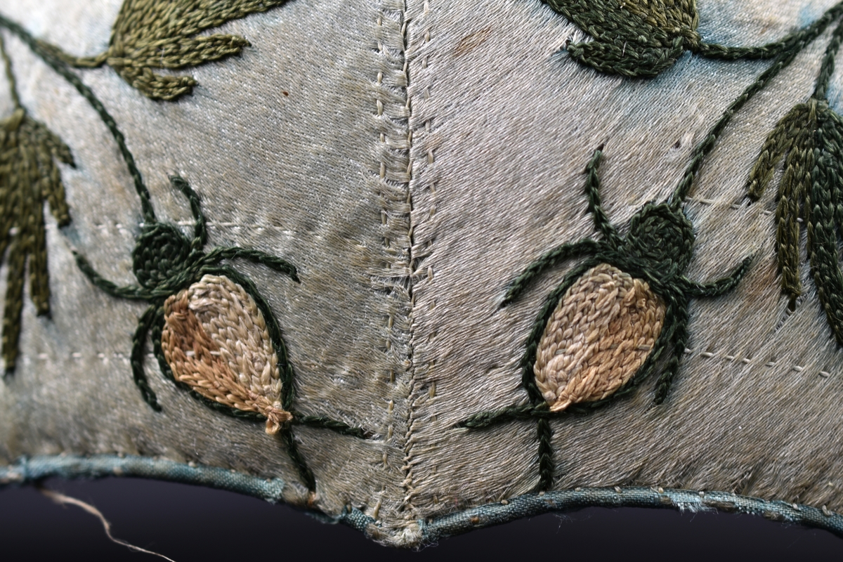 Bindmössa av ljusblått siden dekorerad med broderade blommor och blad. Foder i oblekt linnetyg.