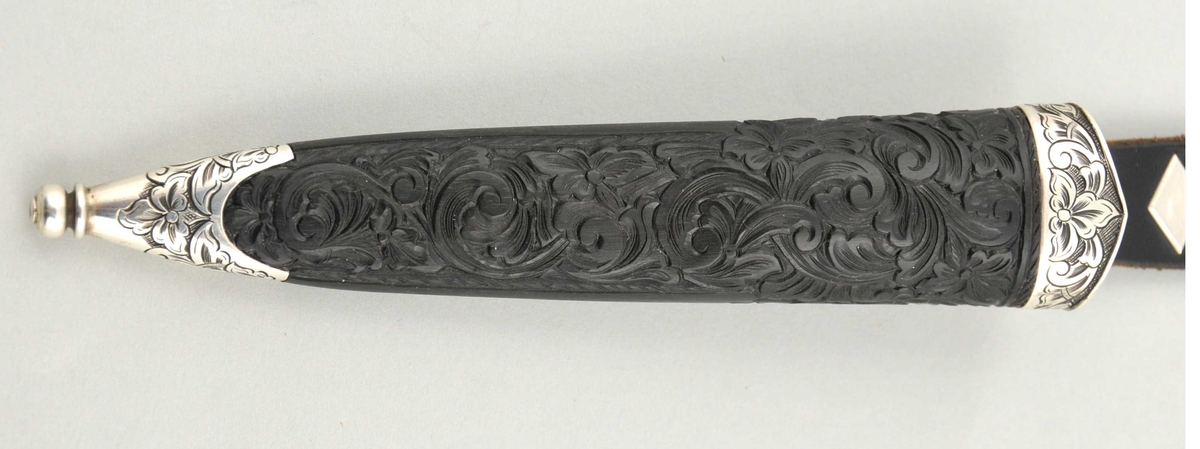 Kniv med utskore skaft og gravera holkar, på toppen ein støypt hund.
Slire av horn, utskoren på begge sider, holkar av sylv som er gravera.