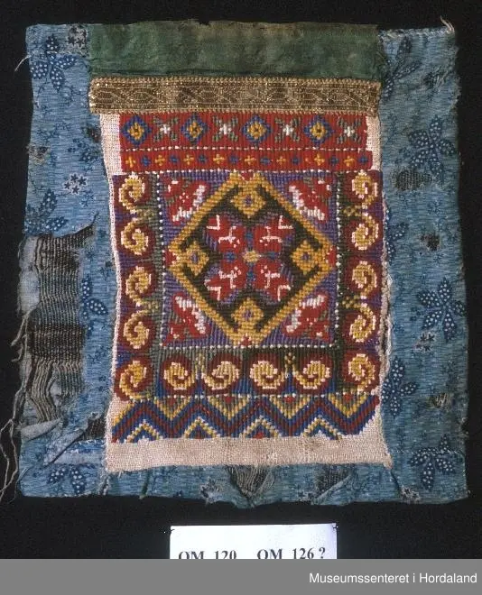 Bringeklut med kross-sting, gullband og grønn silkekvarde. Montert på blått bakstykke med blomstermotiv.