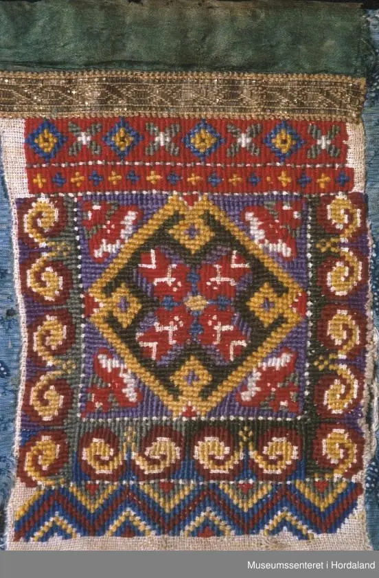 Bringeklut med kross-sting, gullband og grønn silkekvarde. Montert på blått bakstykke med blomstermotiv.