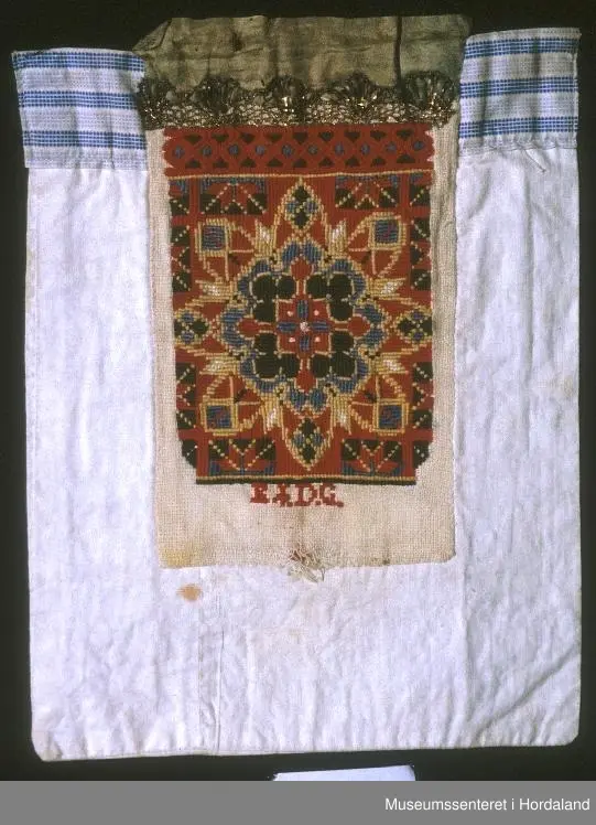 Bringeklut med broderi i fleire farger sydd på eit naturfarga stoff. Breit, grønt silkeband øvst. Gullfarga kronekvarde sydd oppå. Bakstykket er eit dobbelt kvitt stoff med ein kant oppe av eit mønstra stoff i striper, maskinsydd.
Under kronekvarden er det dragesaum. Ellers
kross-sting i broderiet.