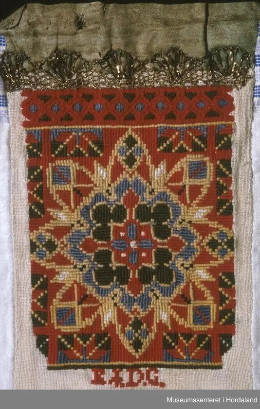 Bringeklut med broderi i fleire farger sydd på eit naturfarga stoff. Breit, grønt silkeband øvst. Gullfarga kronekvarde sydd oppå. Bakstykket er eit dobbelt kvitt stoff med ein kant oppe av eit mønstra stoff i striper, maskinsydd.
Under kronekvarden er det dragesaum. Ellers
kross-sting i broderiet.