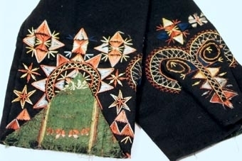 Svarta vantar med broderi i rätlinjig plattsöm och flätsöm med silke i rött, blått, gult. En snibbliknande flik, klädd med grönt siden, är uppvikt mot framsidans nederkant.