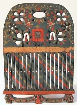 Bandvävsked av trä med genombrutet överstycke med en liten fyrkantig spegelglasbit inom röd ram på mitten. Bandgrinden är målad i gråblått med ornering i rött, gult, svart och vitt.