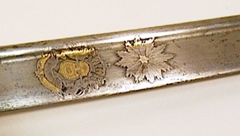 Infanteriofficerssabel. Fästenas beslag är av förgylld mässing och parerplåten är dekorerad med genombrutna ornament. Kaveln är klädd med svärtat fiskskinn, med åtta refflor i vars bottnar tvinnade mässingstrådar anbringats. . På klingans flatsidor finns etsade och delvis förgyllda ornament.