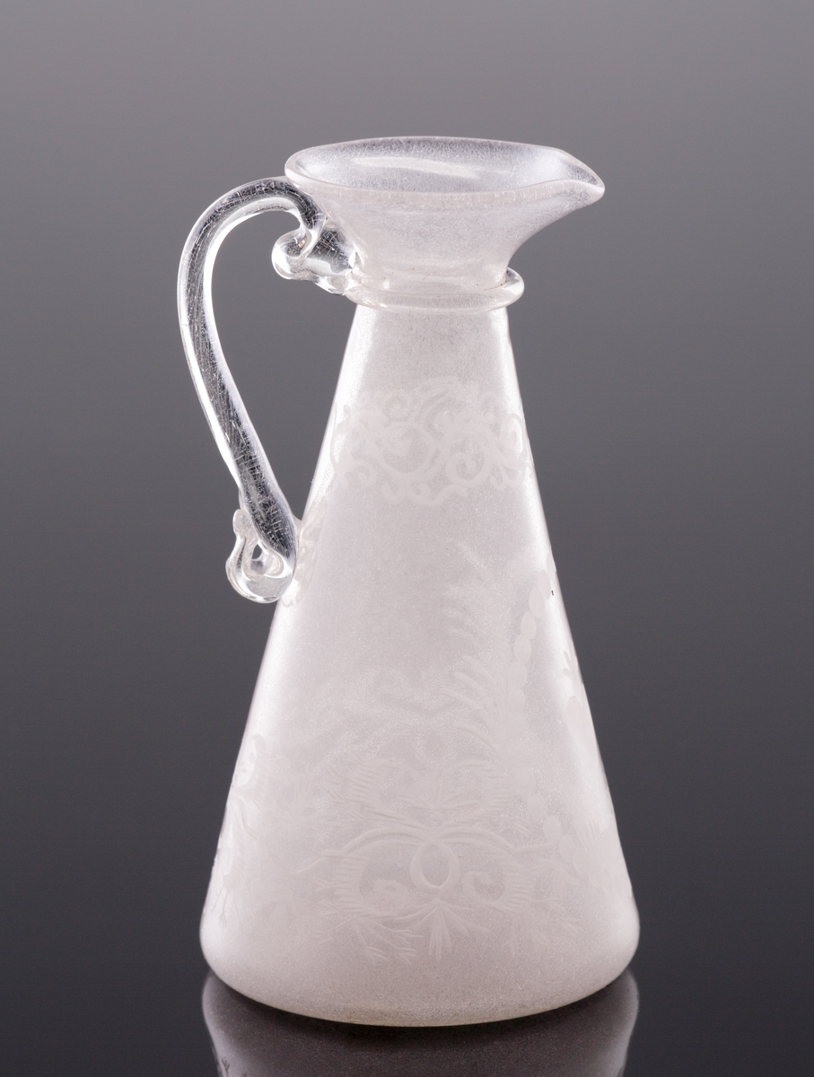 Kanna. 
Konande modell med hänkel och graverad dekor av bladrankor, två brinnande hjärtan m.m.
Mynningen utvikt med en formad pip. Under mynningen en påklippt ring.