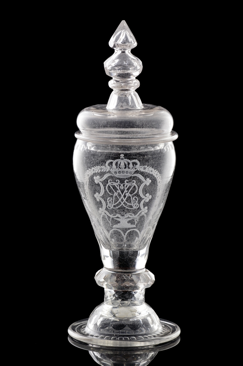 Pokal av glas, med lock. 
Slipat och graverat mönster med kungligt spegelmonogram: "FR"* i kartusch under krona. Invikt kant på foten.

*"FR" (Fredericus Rex= Fredrik I av Sverige, reg. 1720-1751).