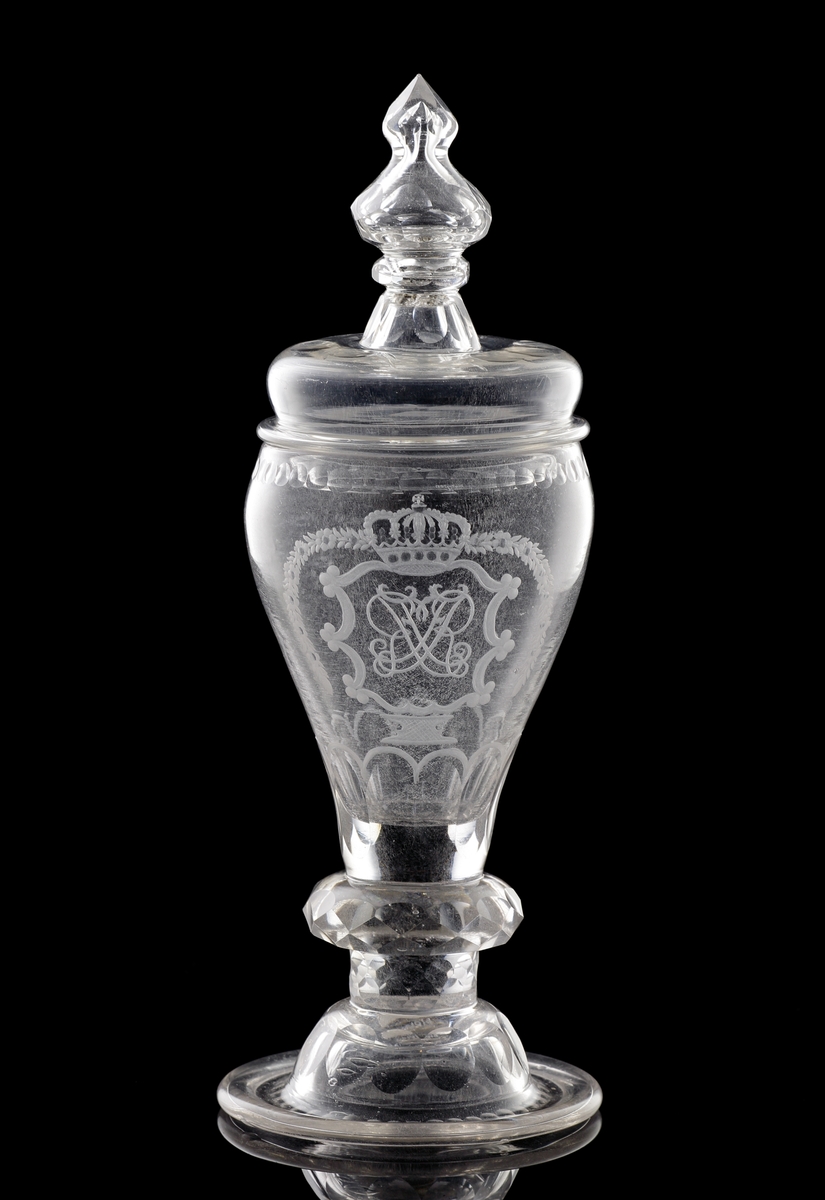 Pokal av glas, med lock. 
Slipat och graverat mönster med kungligt spegelmonogram: "FR"* i kartusch under krona.

*"FR" (Fredericus Rex= Fredrik I av Sverige, reg. 1720-1751).