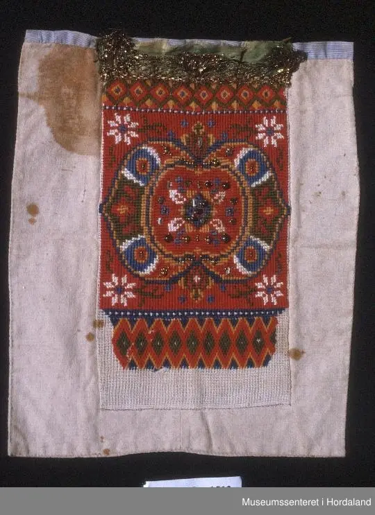 Bringeklut brodert på stramei med kross-sting og dragesaum. Raud med blått, gult, grønt og gull. Festa på eit kvitt stoff. Stoffet har ein stor flekk og nokre små. Oppe på baksida er montert på eit stripete stoff som går eit lite stykke ned på framsida. Grønt silkeband oppe med gullfarga metallkronekvarde sydd utanpå. Store og små perler sydd på broderimønsteret, nokre av desse er knuste.