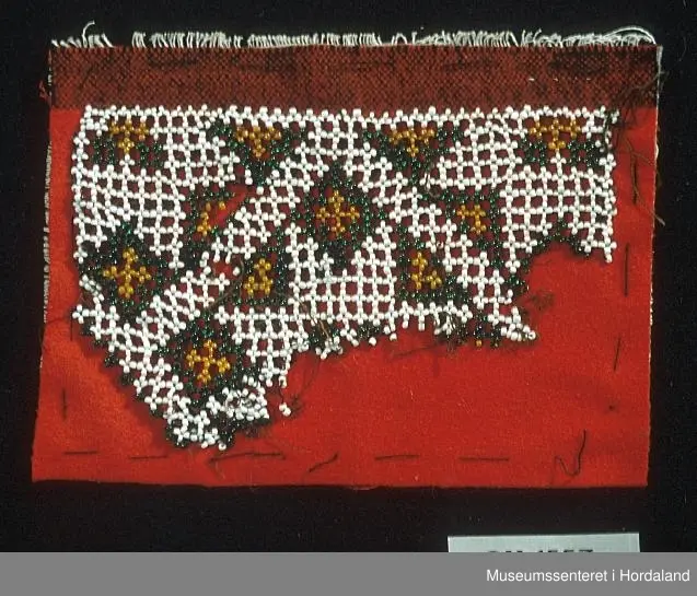 Uferdig bringeklut med perlebroderi i kvitt,gult og grønt på raud bakgrunn.