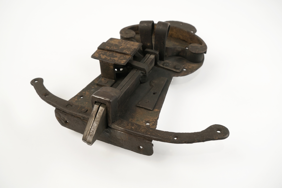 Dørlås av jern. Formet som en person, som står på en meie. Grov låsemekanisme med en hendel som gjør at låsebolten kan skyves til side, og dermed åpne døren.